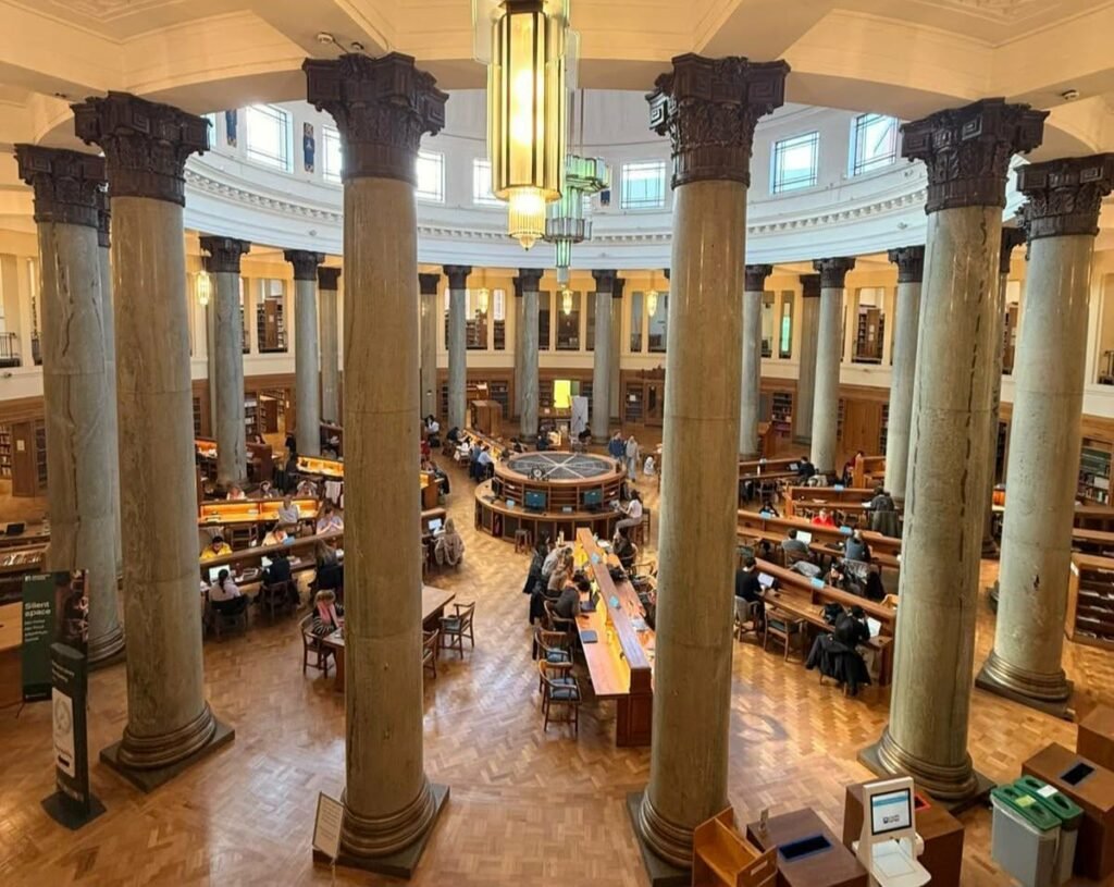 بين الحرف والحدّ: رحلة في متاهة مكتبة بروذرتون "Interior view of the historic Brotherton Library at the University of Leeds, featuring the iconic circular reading room with majestic marble columns, a high domed ceiling, and students seated at classic wooden study tables surrounded by tiers of bookshelves."