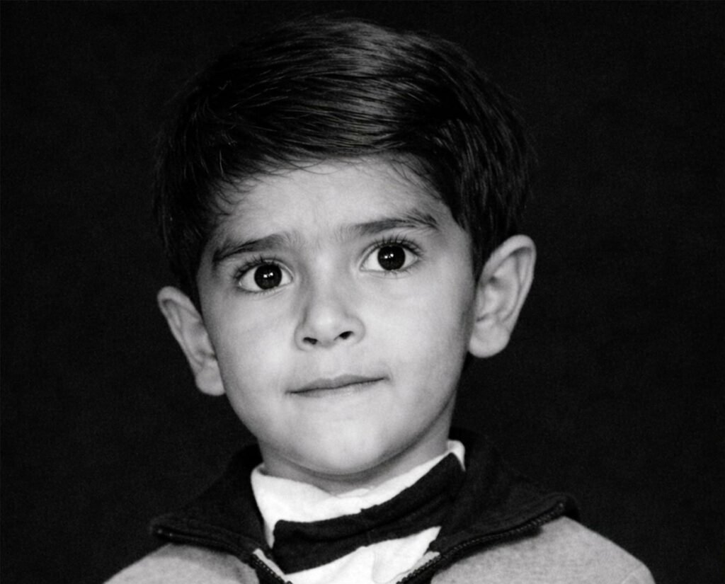 A vintage black and white studio portrait of a young boy with a serious expression, looking directly at the camera. He is wearing a shirt with a small bow tie under a zippered jacket. The photo features a dark, minimalist background typical of 1960s Iraqi photography studios.