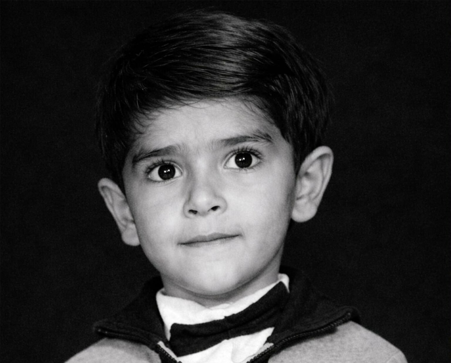 A vintage black and white studio portrait of a young boy with a serious expression, looking directly at the camera. He is wearing a shirt with a small bow tie under a zippered jacket. The photo features a dark, minimalist background typical of 1960s Iraqi photography studios.