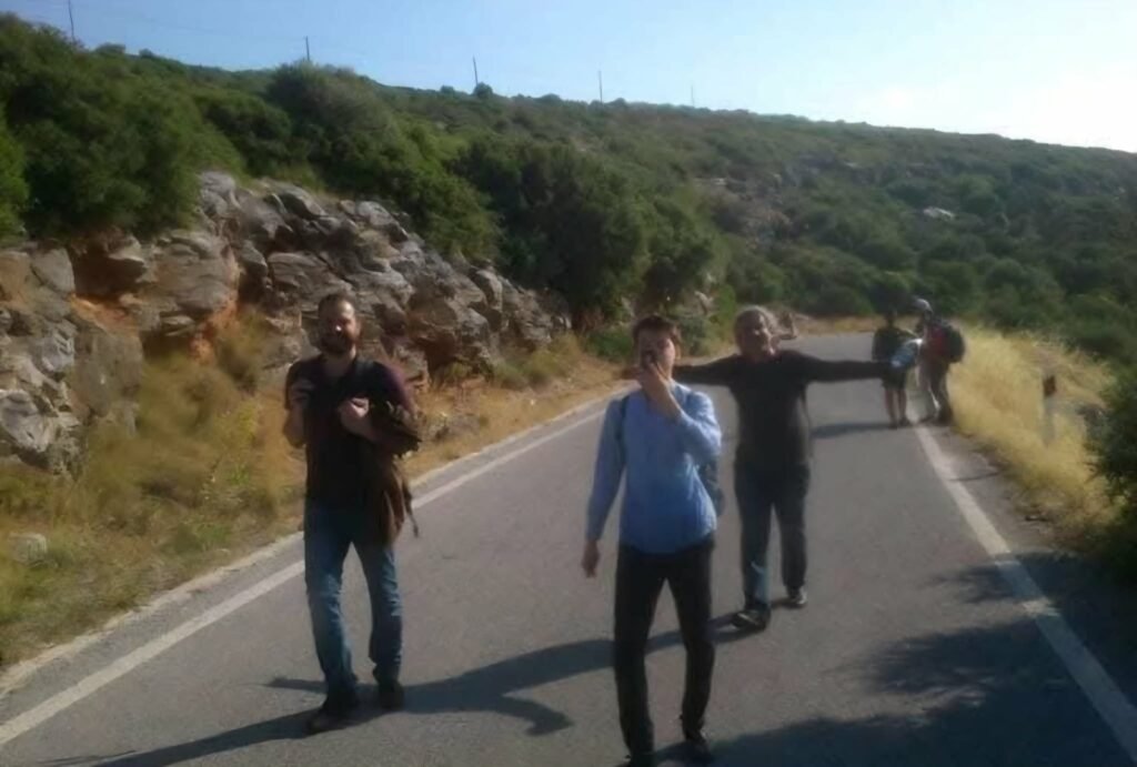 سترة نجاة برتقالية مثقوبة في الطريق إلى إيثاكا! Syrian refugees, wearing orange life jackets, walk on a steep asphalt road downhill after landing on the coast of Samos Island, Greece, in Autumn 2015. Arid hills and small buildings are visible under bright sunlight, capturing a moment of survival and uncertainty during the migration crisis.