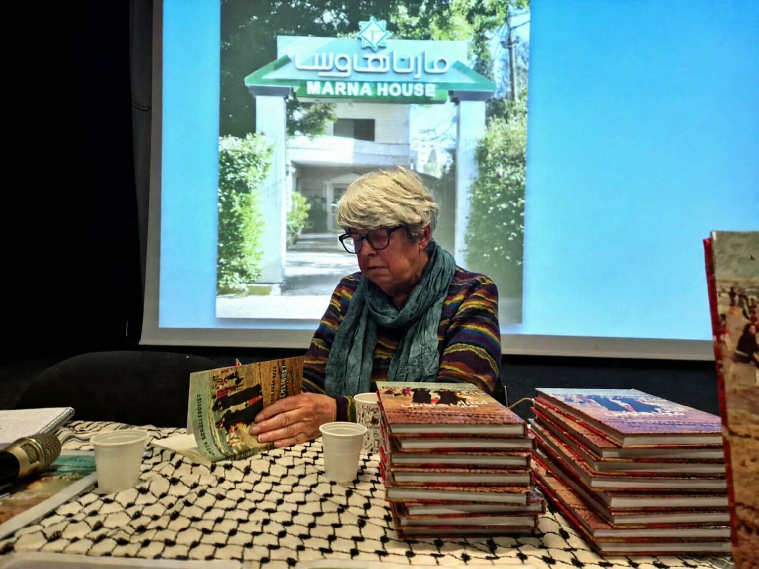 Swedish author Lotta Schullerqvist sits at a book signing event for the Arabic edition of her book 'Secrets of Marna House'. She is behind a table with copies of the book, with a backdrop featuring the book's title in Arabic and an illustration of a traditional Palestinian house, symbolizing Gaza's memory and heritage.