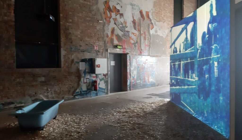 An installation view from the photography exhibition, showing a minimalist gallery setup with multi-layered visual displays on the walls, a wooden structures element, and soft artistic lighting creating shadows in a concrete industrial space.