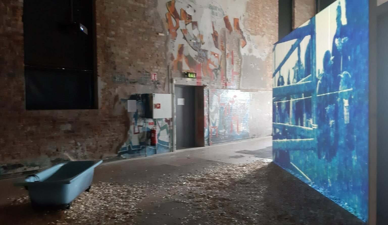 An installation view from the photography exhibition, showing a minimalist gallery setup with multi-layered visual displays on the walls, a wooden structures element, and soft artistic lighting creating shadows in a concrete industrial space.