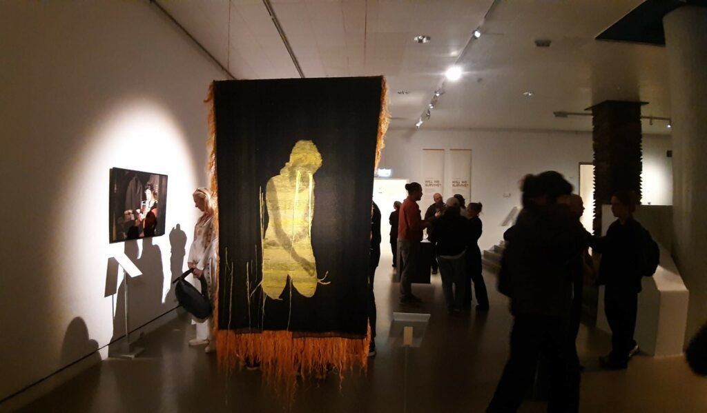 "Installation view of the 'We Will Survive' exhibition at Röhsska Museum, featuring a large black textile with a golden silhouette of a kneeling figure, surrounded by visitors in a dimly lit gallery setting."