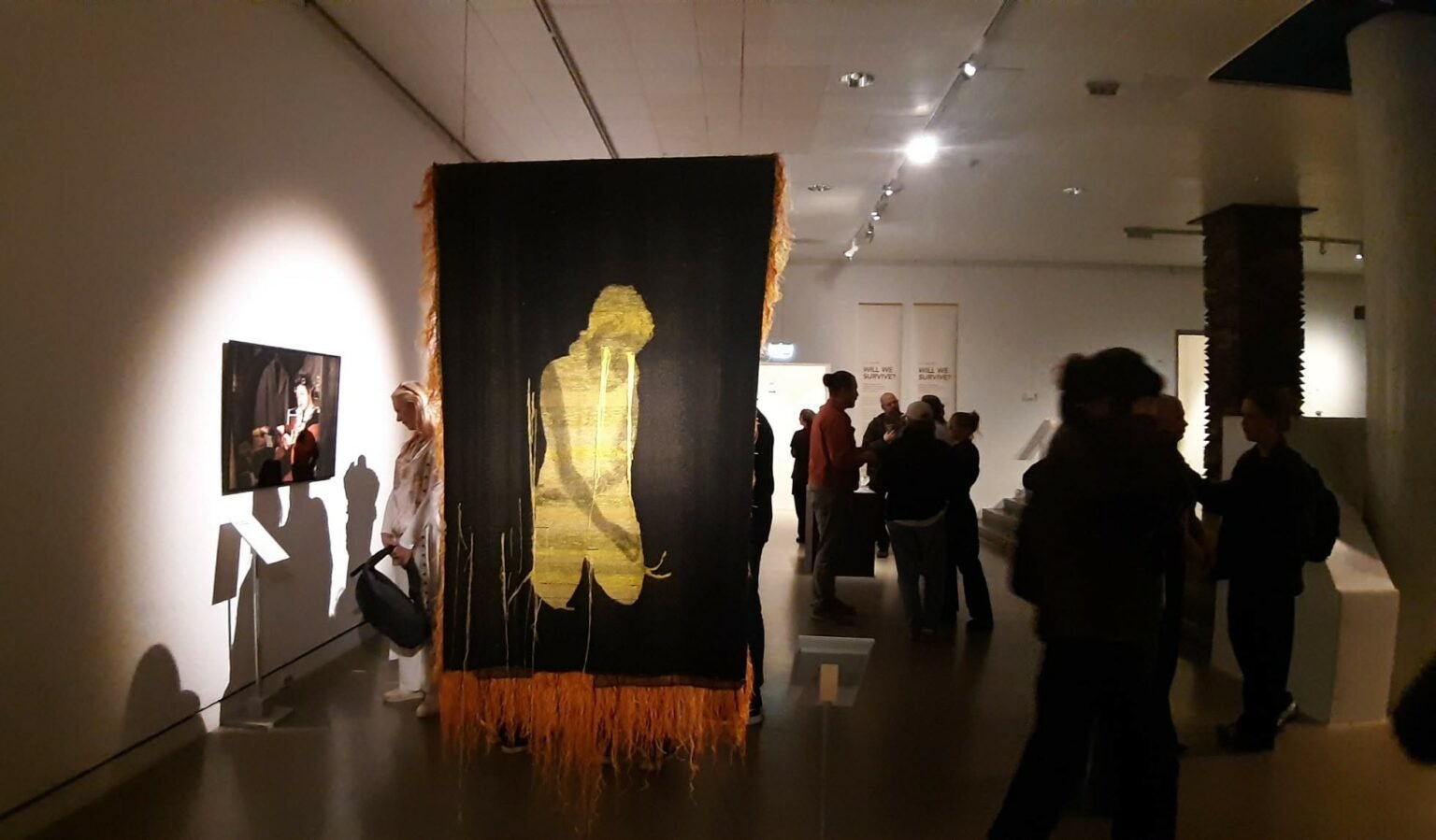 "Installation view of the 'We Will Survive' exhibition at Röhsska Museum, featuring a large black textile with a golden silhouette of a kneeling figure, surrounded by visitors in a dimly lit gallery setting."