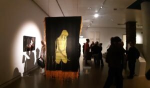 "Installation view of the 'We Will Survive' exhibition at Röhsska Museum, featuring a large black textile with a golden silhouette of a kneeling figure, surrounded by visitors in a dimly lit gallery setting."