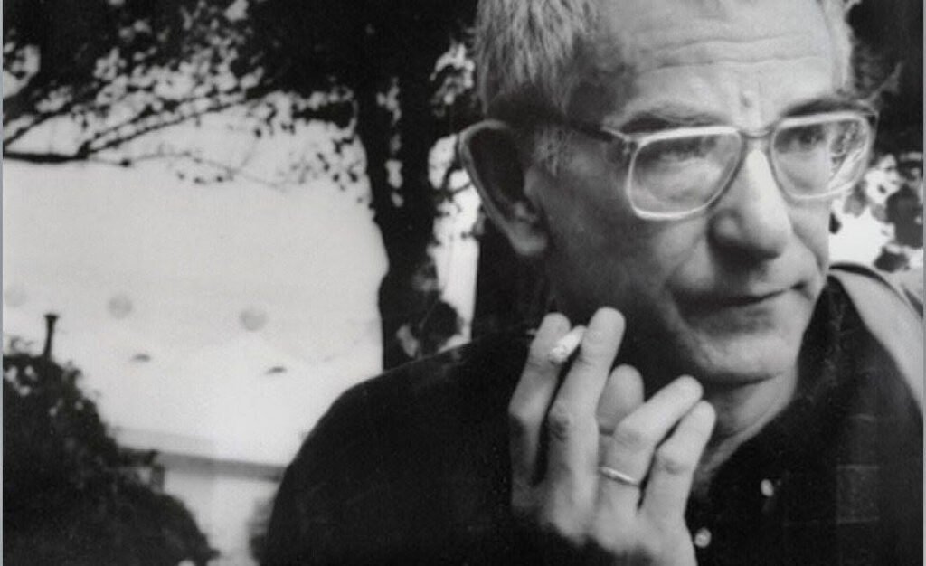 "A black and white close-up portrait of the Polish film director Krzysztof Kieslowski. He is looking slightly to the right with a pensive expression, wearing wire-rimmed eyeglasses. He is holding a burning cigarette between his fingers close to his chin, with a soft-focus background of trees and a building."