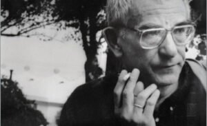 "A black and white close-up portrait of the Polish film director Krzysztof Kieslowski. He is looking slightly to the right with a pensive expression, wearing wire-rimmed eyeglasses. He is holding a burning cigarette between his fingers close to his chin, with a soft-focus background of trees and a building."