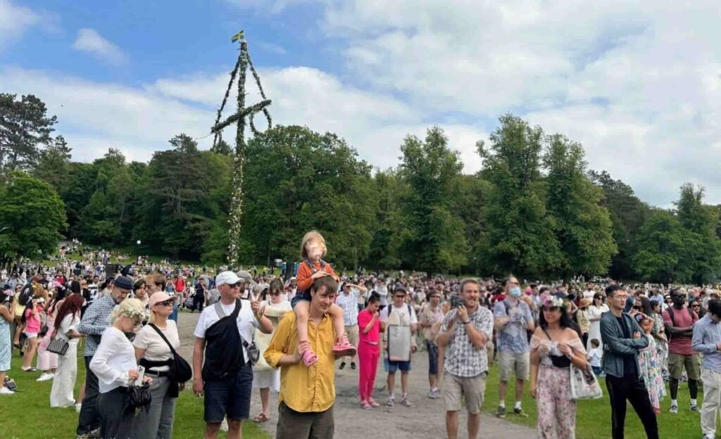 Large crowd of people gathered in Slottsskogen park in Gothenburg, Sweden, celebrating Midsommar (Midsummer) with a traditional decorated maypole standing under a blue sky with clouds.
