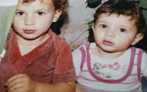 "A vintage, slightly faded color photograph from a family album showing two young children sitting close together. On the left, an older child with curly brown hair wearing a reddish-brown velvet short-sleeved shirt looks towards the camera. On the right, a younger baby with short dark hair, wearing a white sleeveless top with pink stripes and a small cartoon print, gazes forward with a curious expression. The background shows hints of green indoor plant leaves, capturing a nostalgic moment from the past."