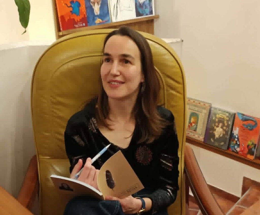 Exclusive portrait of the Bulgarian poet Sybila Alexova holding a pen and her poetry book, sitting on an armchair for Alcommunita website.