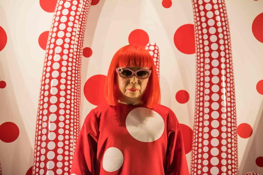 A wax statue or portrait of Japanese artist Yayoi Kusama with bright red hair, wearing a red polka-dot dress and sunglasses, standing in front of a white wall covered in red polka dots and large patterned sculptures.