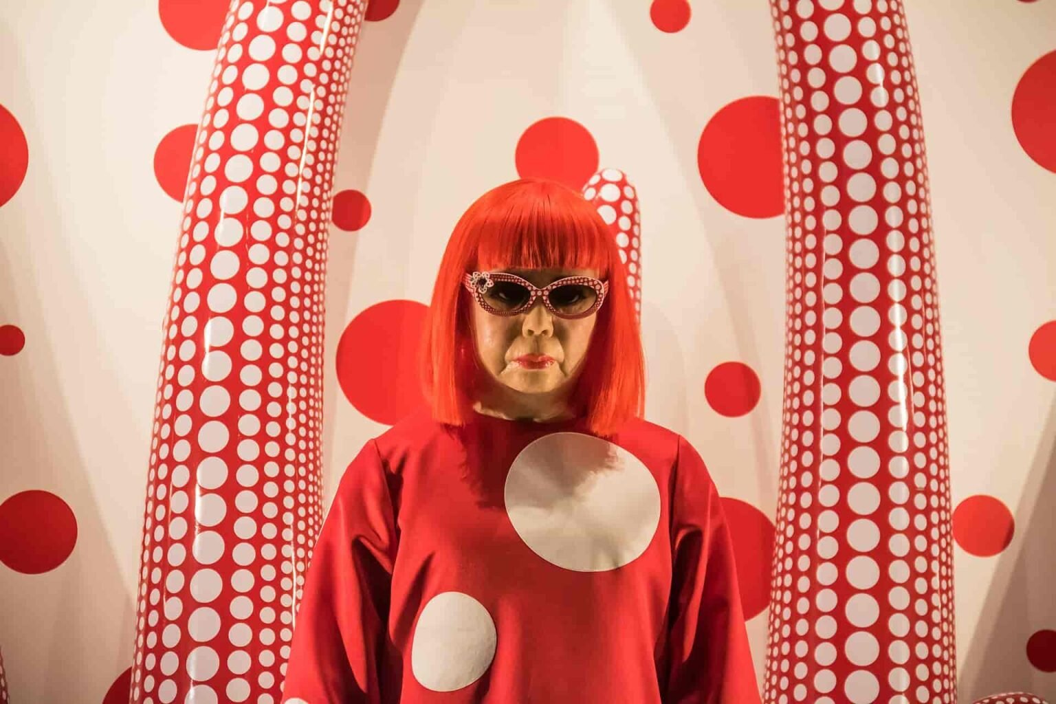 A wax statue or portrait of Japanese artist Yayoi Kusama with bright red hair, wearing a red polka-dot dress and sunglasses, standing in front of a white wall covered in red polka dots and large patterned sculptures.