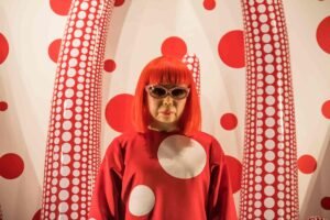 A wax statue or portrait of Japanese artist Yayoi Kusama with bright red hair, wearing a red polka-dot dress and sunglasses, standing in front of a white wall covered in red polka dots and large patterned sculptures.
