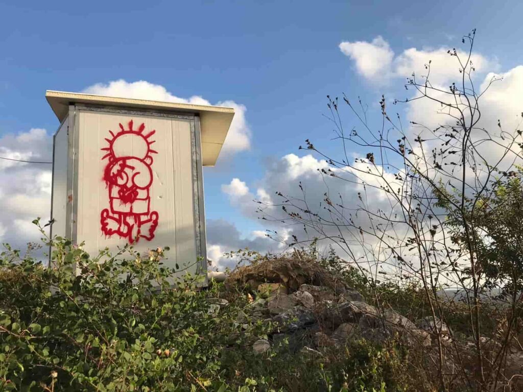 Red spray-painted graffiti of the iconic Palestinian character Handala on a white structure among greenery in Iqrit village, Palestine.