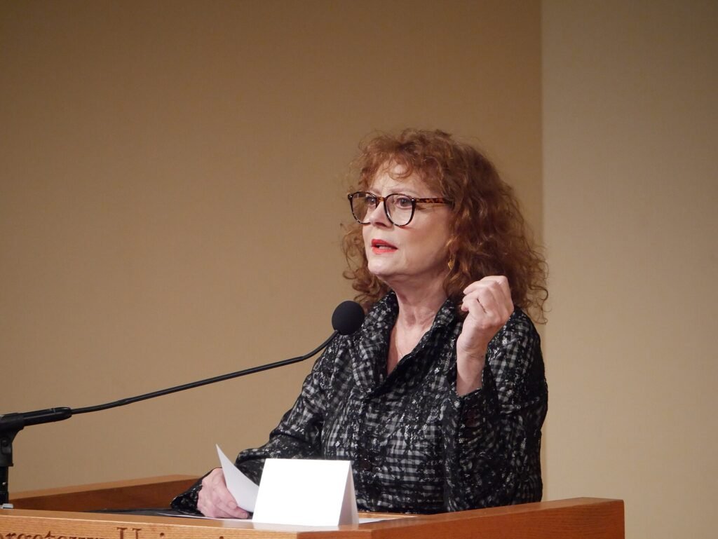 American actress Susan Sarandon wearing glasses speaking into a microphone at a press conference podium.