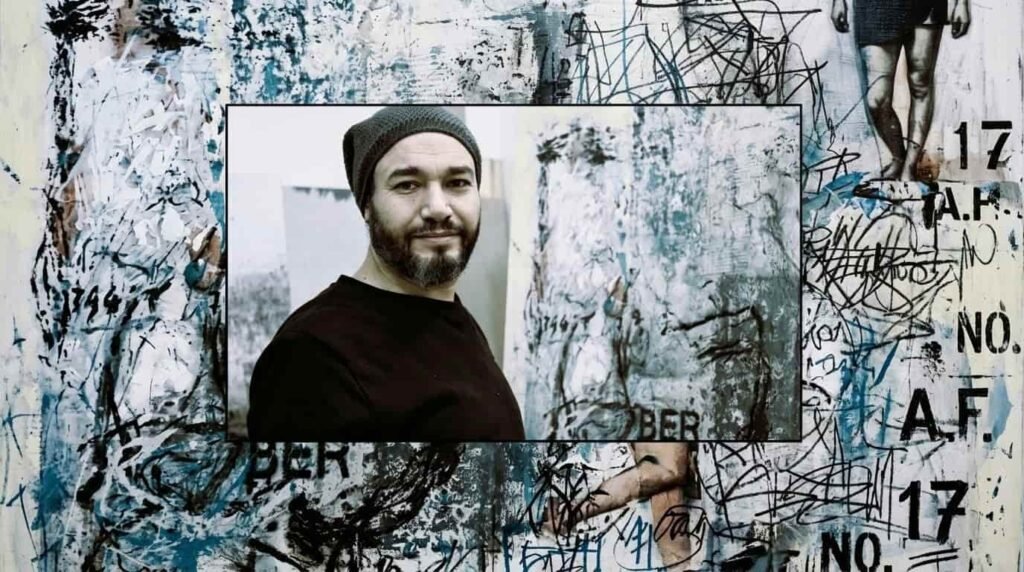A portrait of Syrian artist Abdelkareem Majdal Al-Beik standing next to a textured, weathered wall installation bearing traces and inscriptions in his studio.