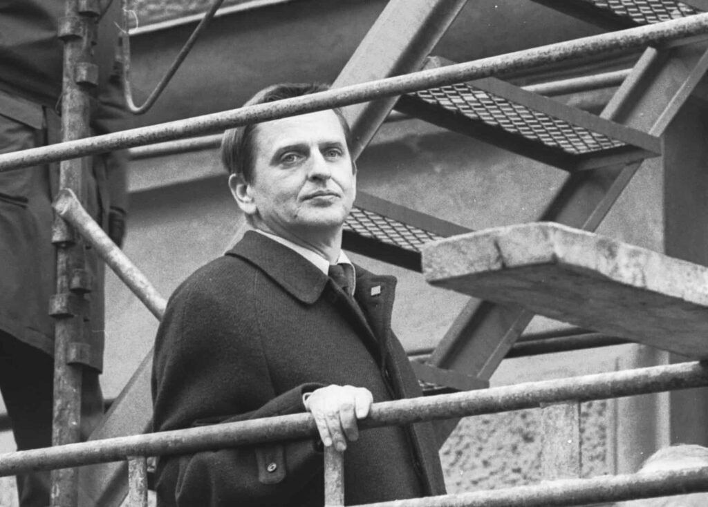 Black and white photograph of former Swedish Prime Minister Olof Palme standing near a metal structure at a May Day rally in Stockholm.
